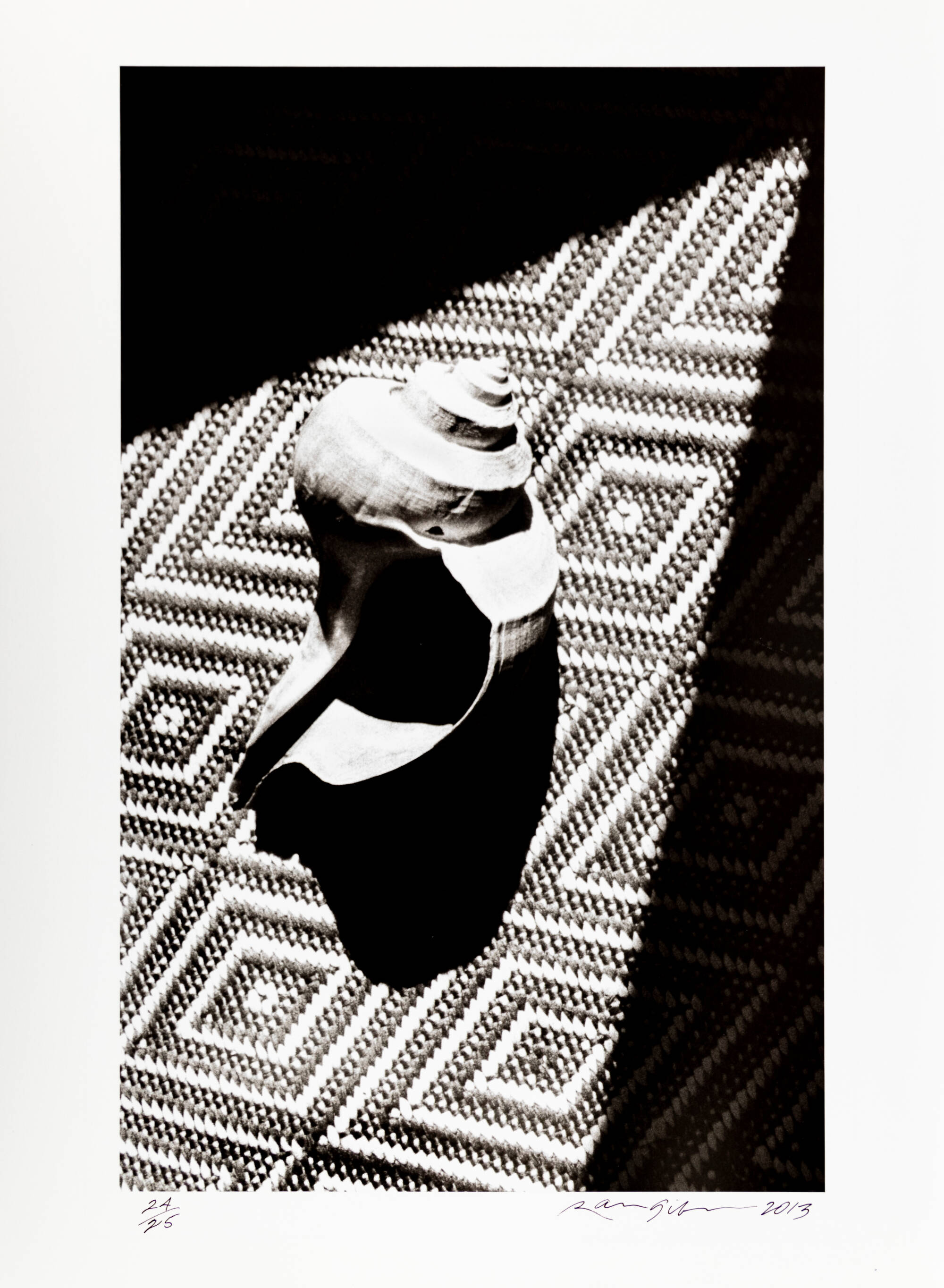 A black and white photo of a shell. The shell sits on a carpet with a diamond-shaped pattern on it.